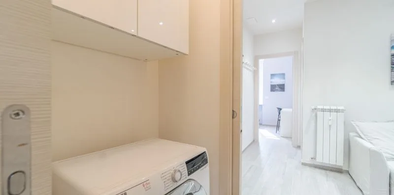 A bright bedroom with a bed, washing machine, and hallway visible.