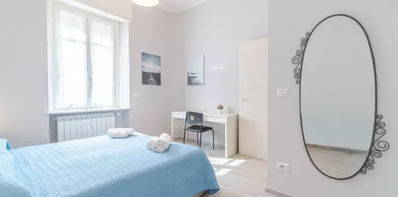 A bedroom with a blue bedspread, a window, and a decorative wall mirror.
