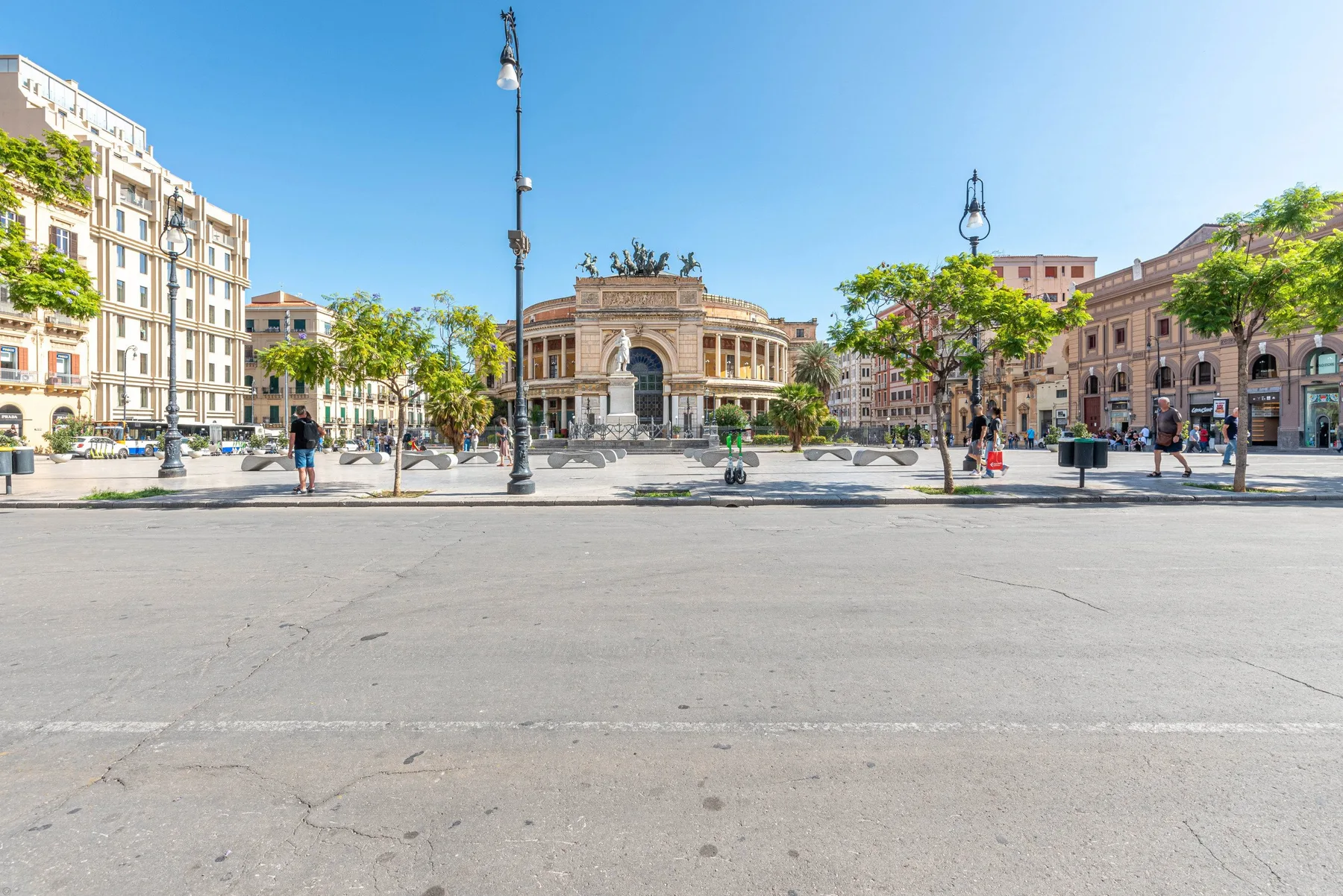 Una plaza pavimentada da a un gran edificio con arcos, árboles y farolas.