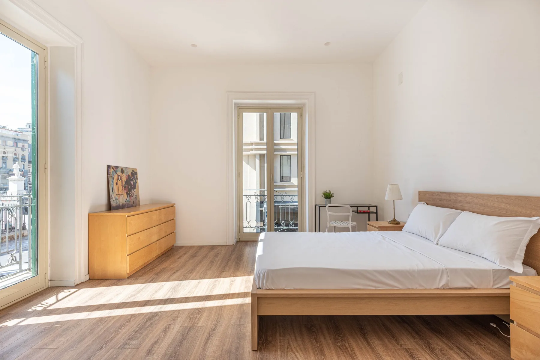 Un dormitorio luminoso con una cama, una cómoda de madera y ventanas grandes.