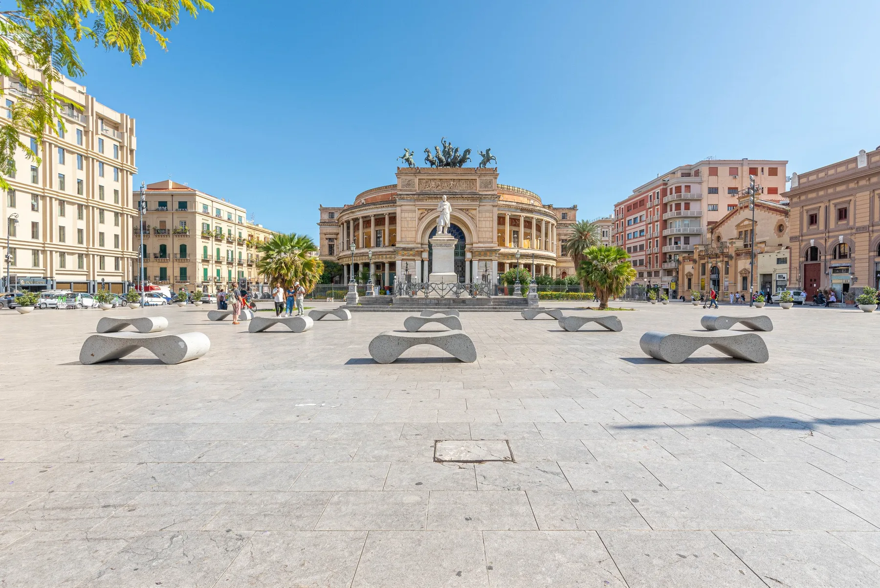 Una plaza soleada con asientos escultóricos y un edificio ornamentado al fondo.