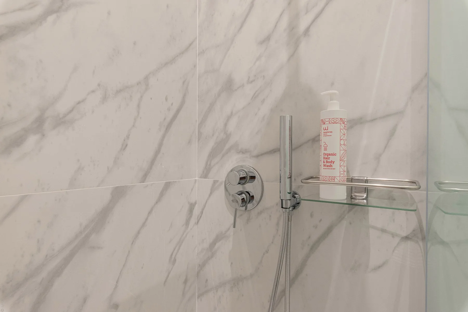 Una ducha con controles cromados y un frasco sobre una repisa de vidrio.