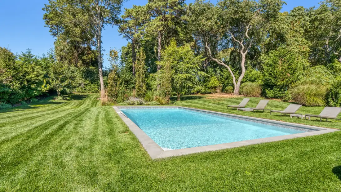 Una piscina está junto a un césped bien cortado y árboles frondosos.