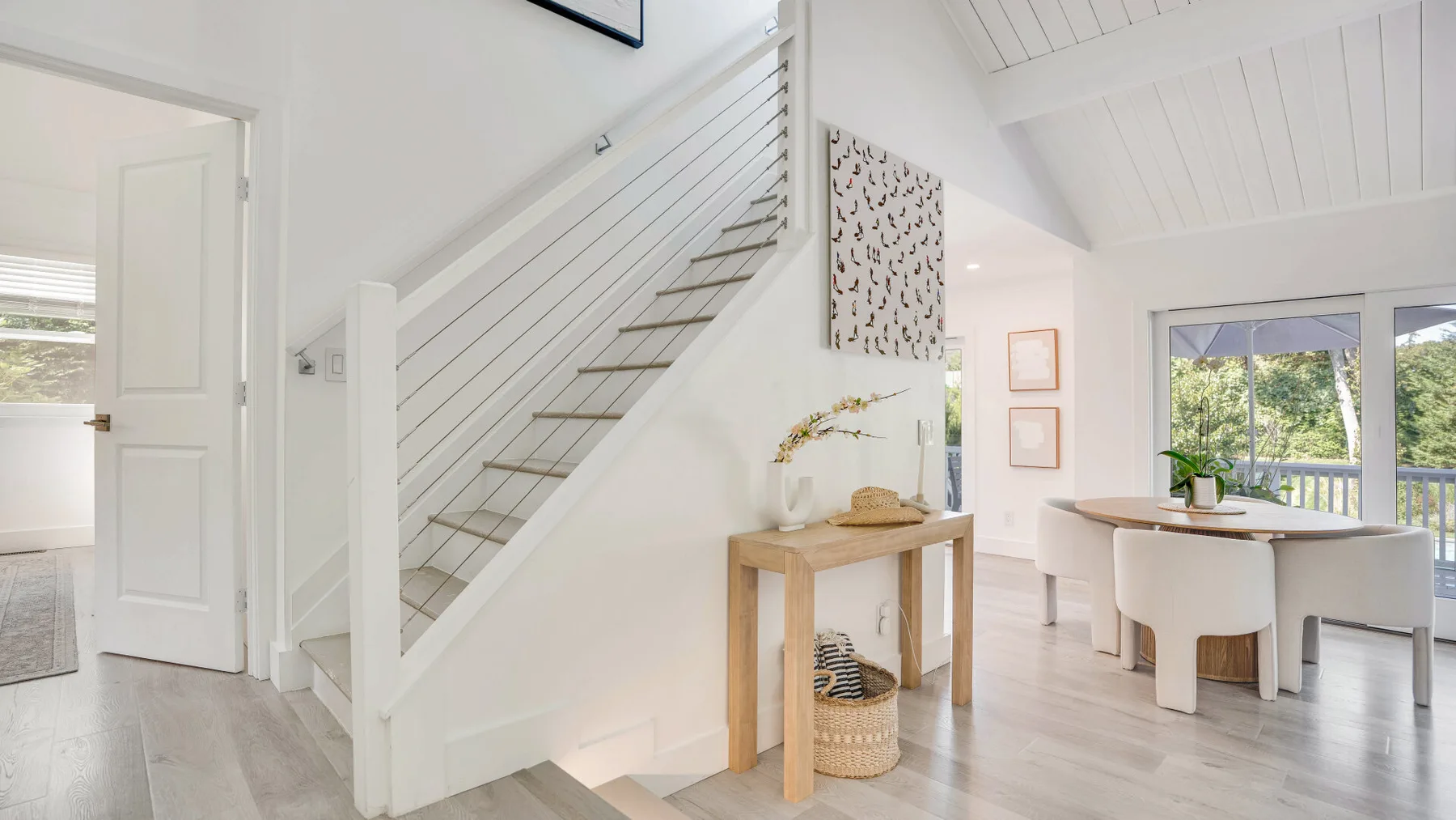 Un comedor luminoso está junto a una escalera blanca, con una mesa y grandes ventanas.