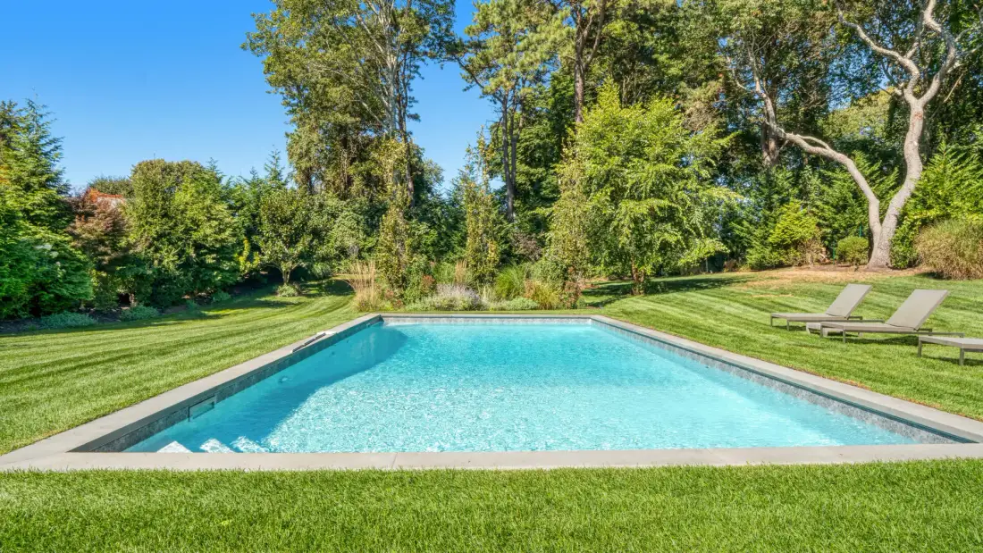 Una piscina al aire libre está en un jardín con césped rodeado de árboles.