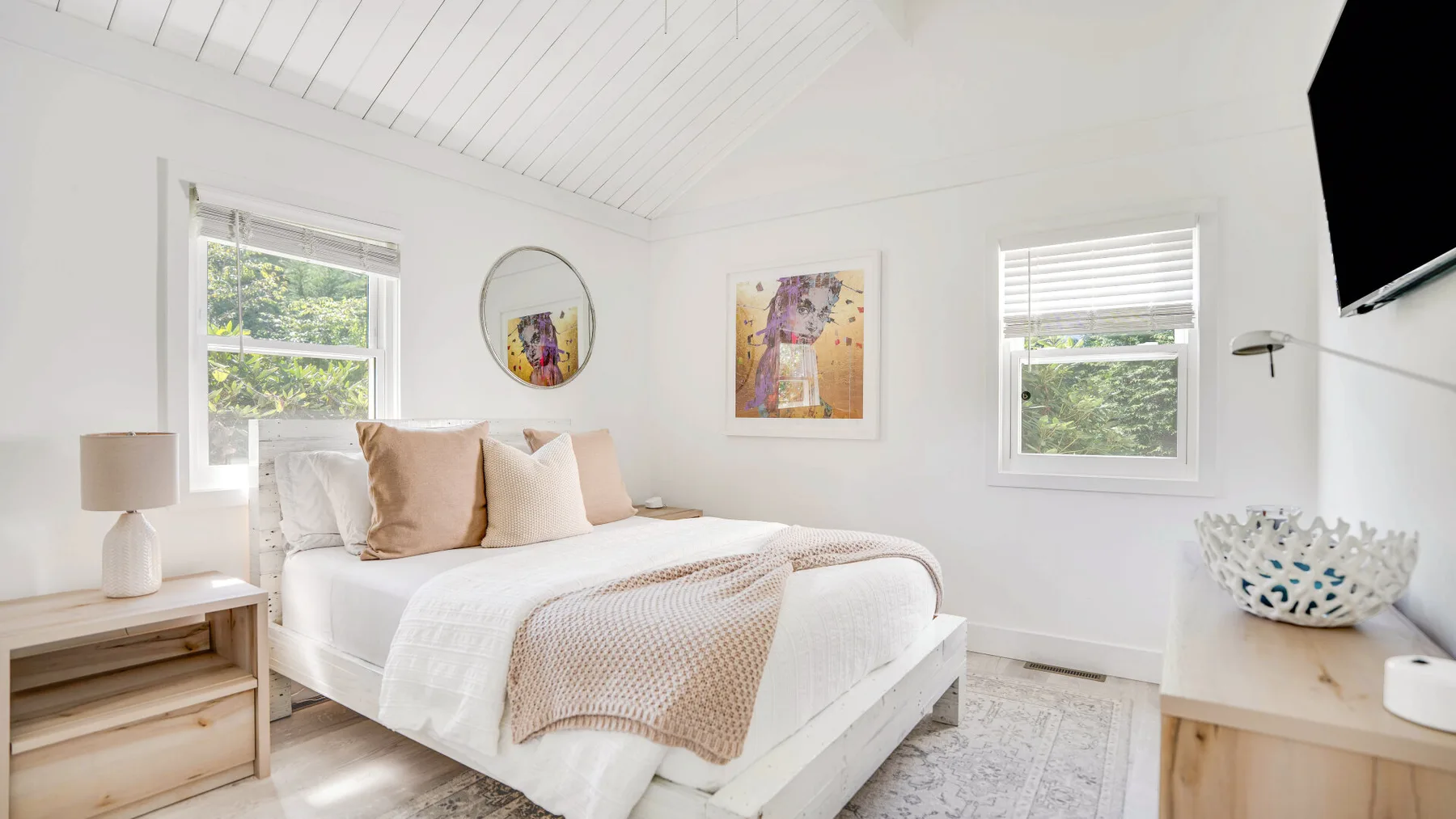 Un dormitorio luminoso con una cama, obras enmarcadas y ventanas con vistas a la vegetación.