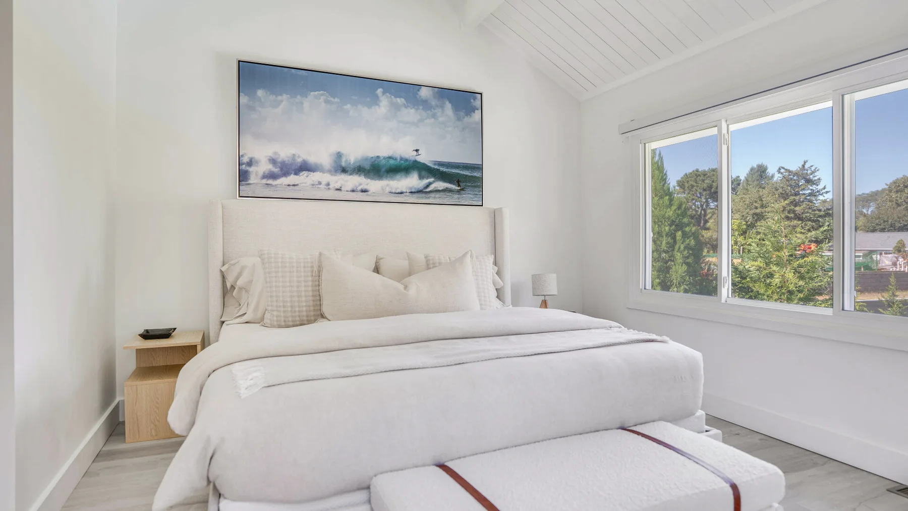 Un dormitorio luminoso con una cama bajo un cuadro del océano y grandes ventanas.