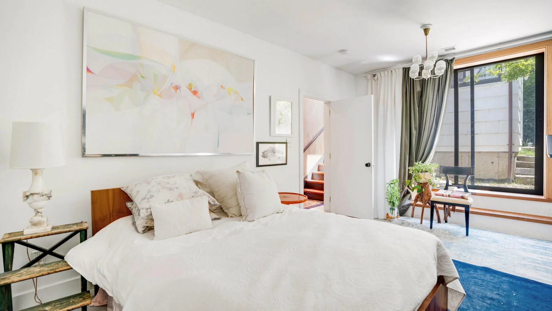 Un dormitorio luminoso con una cama grande, arte abstracto y una ventana con vistas a un patio.