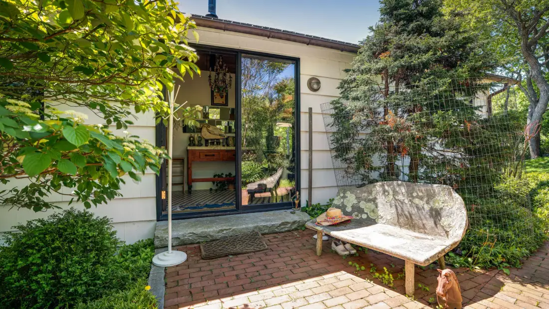 Un patio junto a una puerta de vidrio, con un banco y vegetación alrededor.
