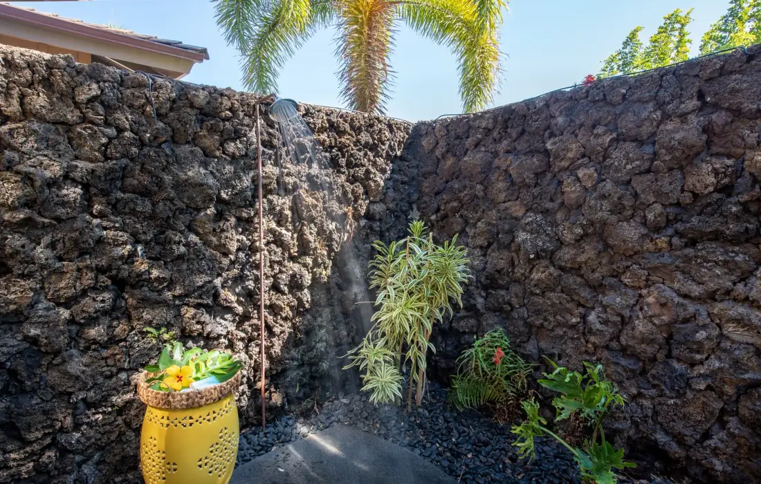 An outdoor shower stands beside a dark stone wall and leafy plants.