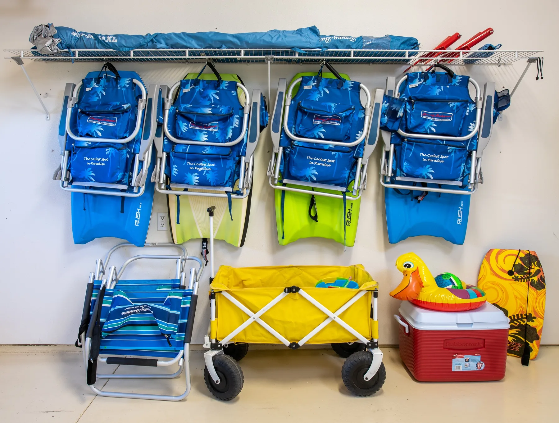 Beach gear is stored on a wall rack above folded chairs, a wagon, and a cooler.