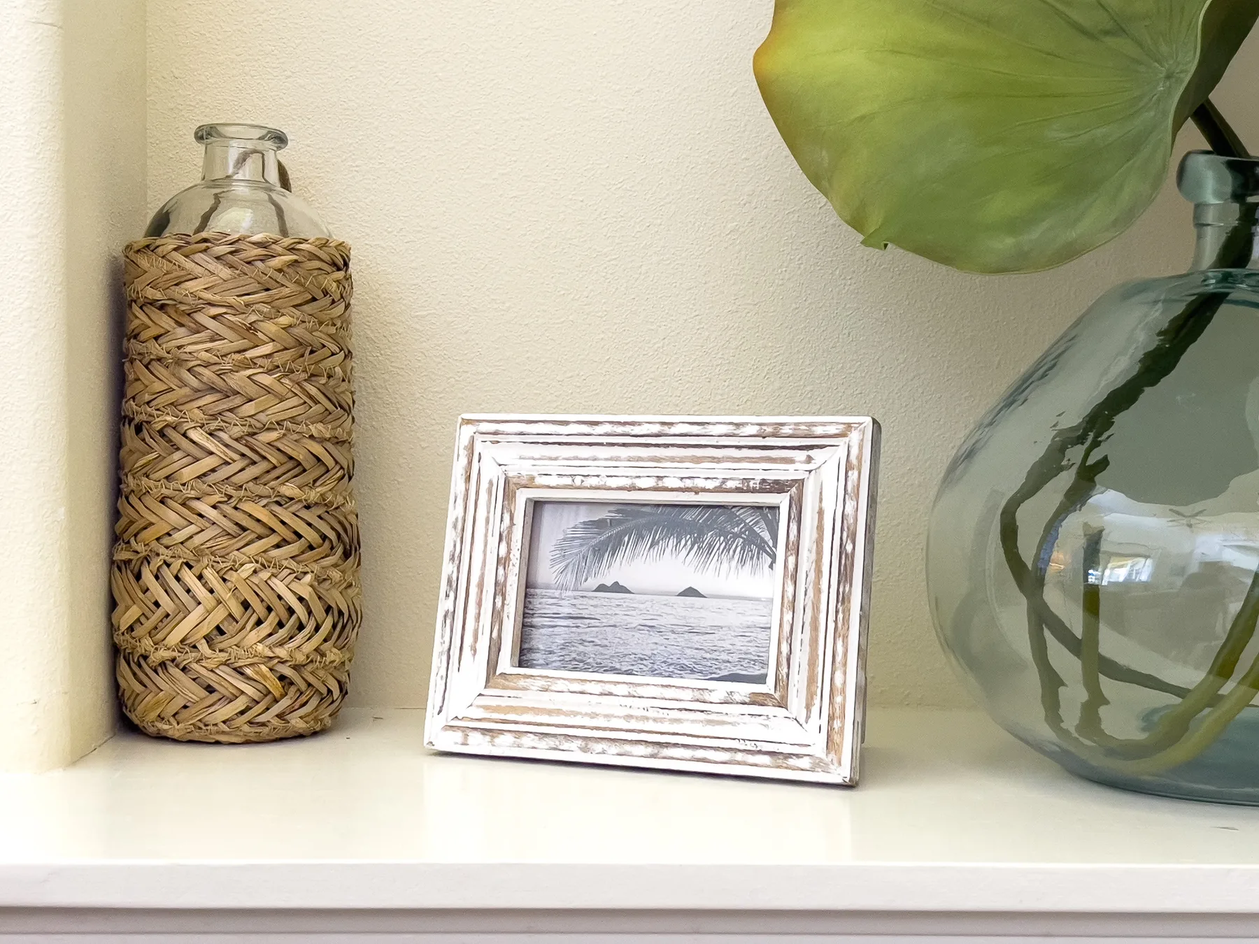 A framed beach photograph sits on a shelf beside decorative glass bottles and a large leaf.