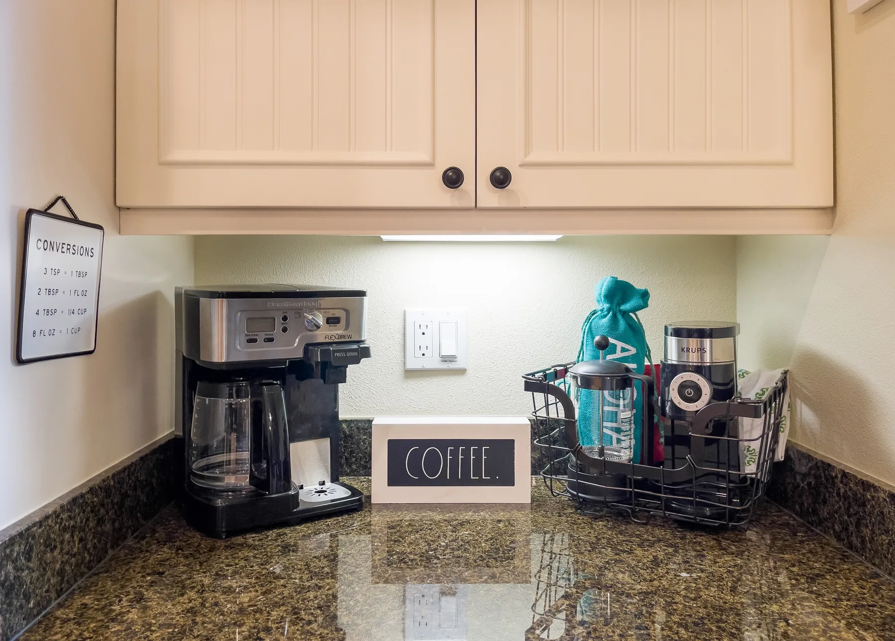 A coffee station sits on a countertop beneath overhead cabinets.