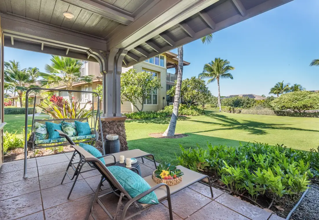 A covered patio with seating overlooks a landscaped lawn and palm trees.