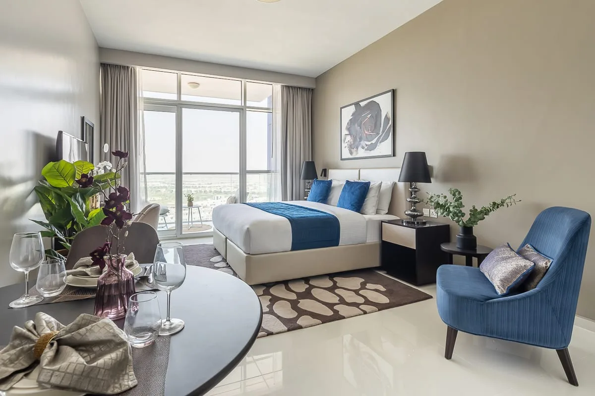 Un dormitorio luminoso con una cama tendida y asientos azules junto a grandes ventanas.