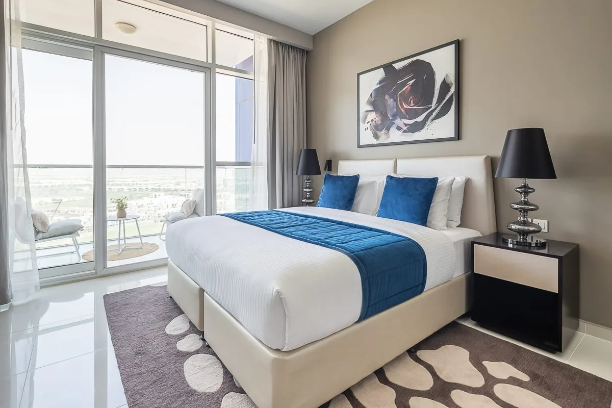 A bright bedroom has a neatly made bed with blue bedding beside large windows.