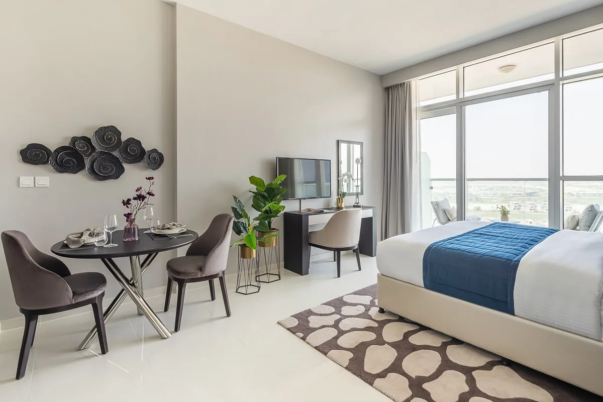 Un dormitorio luminoso con una cama, una pequeña mesa de comedor y grandes ventanas.