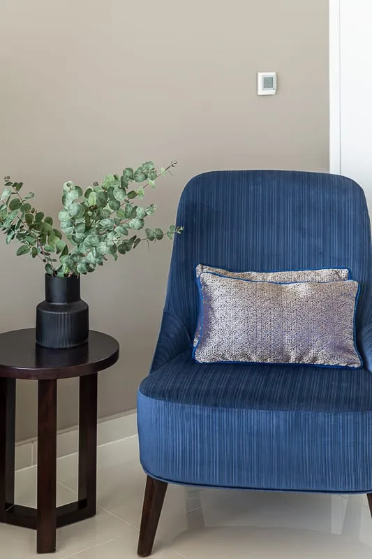 A blue upholstered seat with a decorative cushion sits beside a small round table and leafy plant.