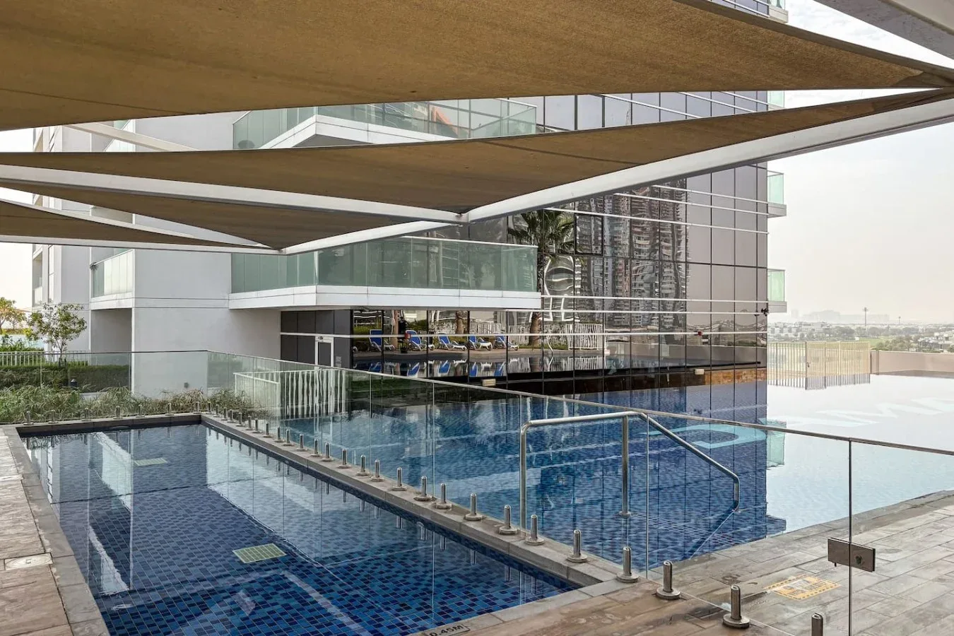 Una piscina exterior está junto a un edificio con fachada de vidrio y toldos de sombra.