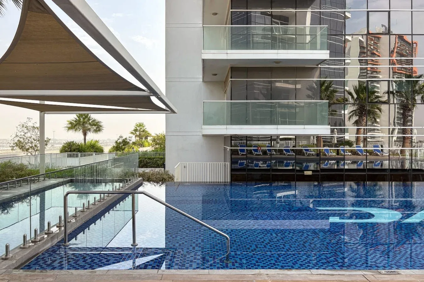 Una piscina al aire libre junto a un edificio moderno y una zona de descanso sombreada.