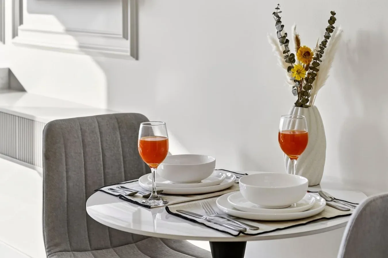 Una mesa está puesta con vajilla blanca, bebidas anaranjadas y un jarrón con flores.