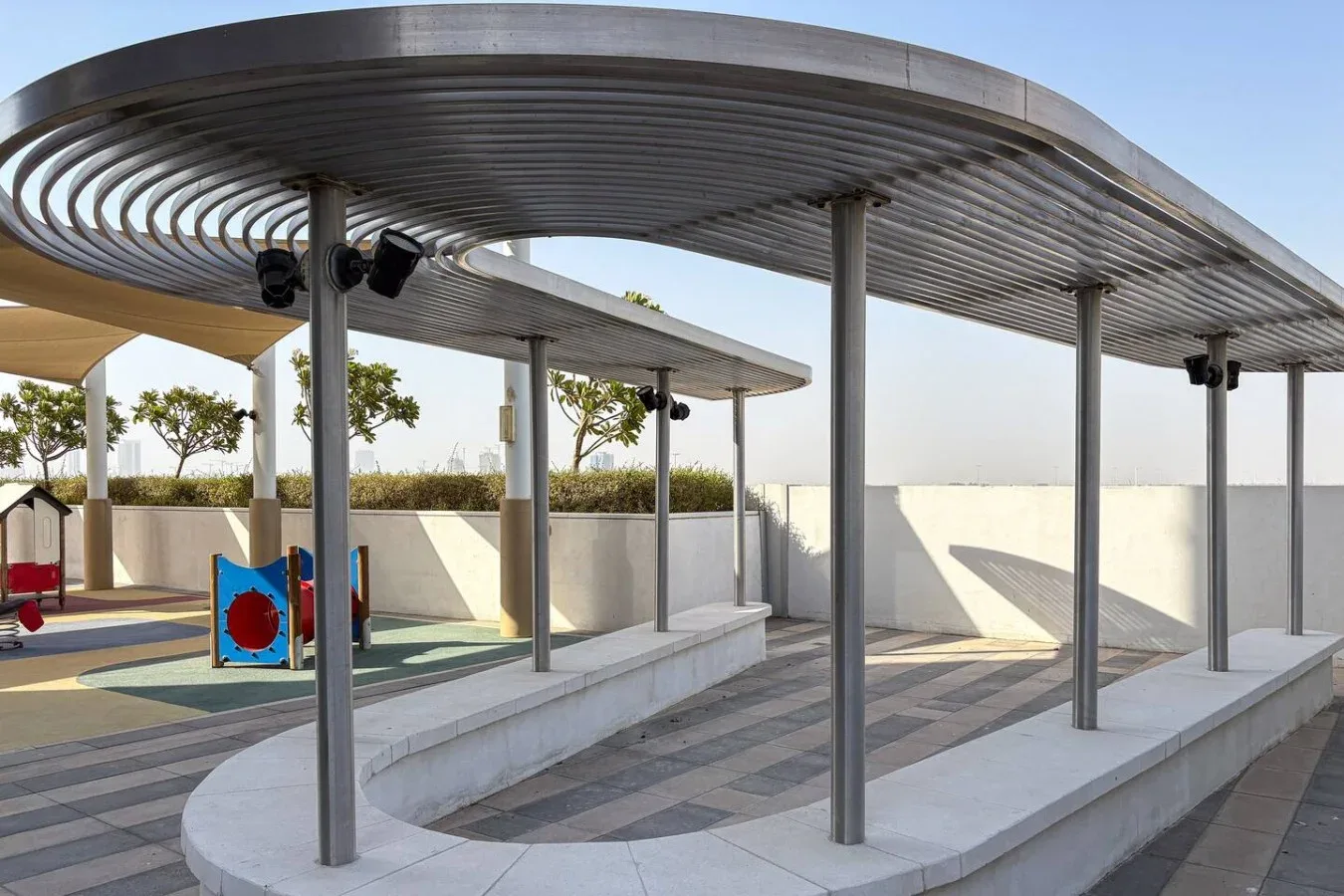 Una zona exterior pavimentada con toldos metálicos curvos y una pequeña estructura de juego.