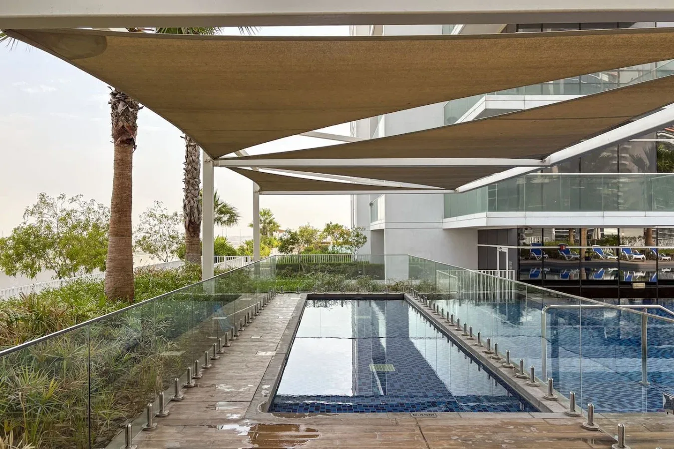Una piscina exterior estrecha bordea un edificio moderno de vidrio bajo toldos de sombra.