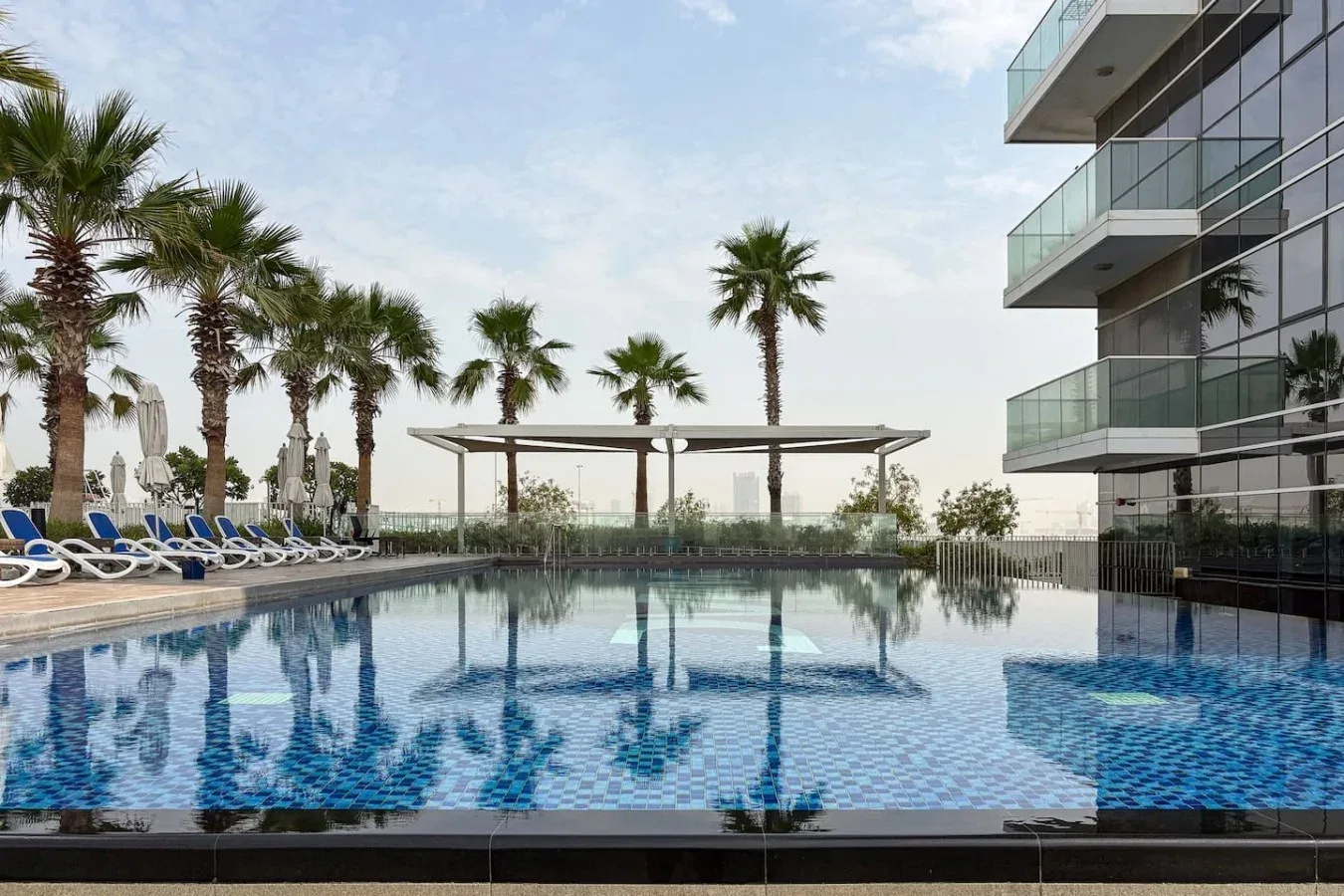 Una piscina exterior junto a un edificio moderno, con palmeras y tumbonas.