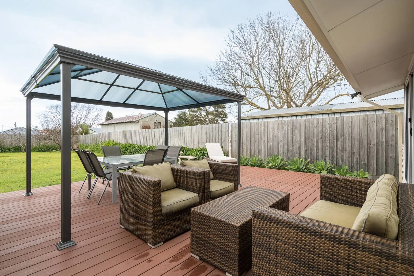 Un patio al aire libre tiene asientos y una mesa de comedor bajo una pérgola cubierta.