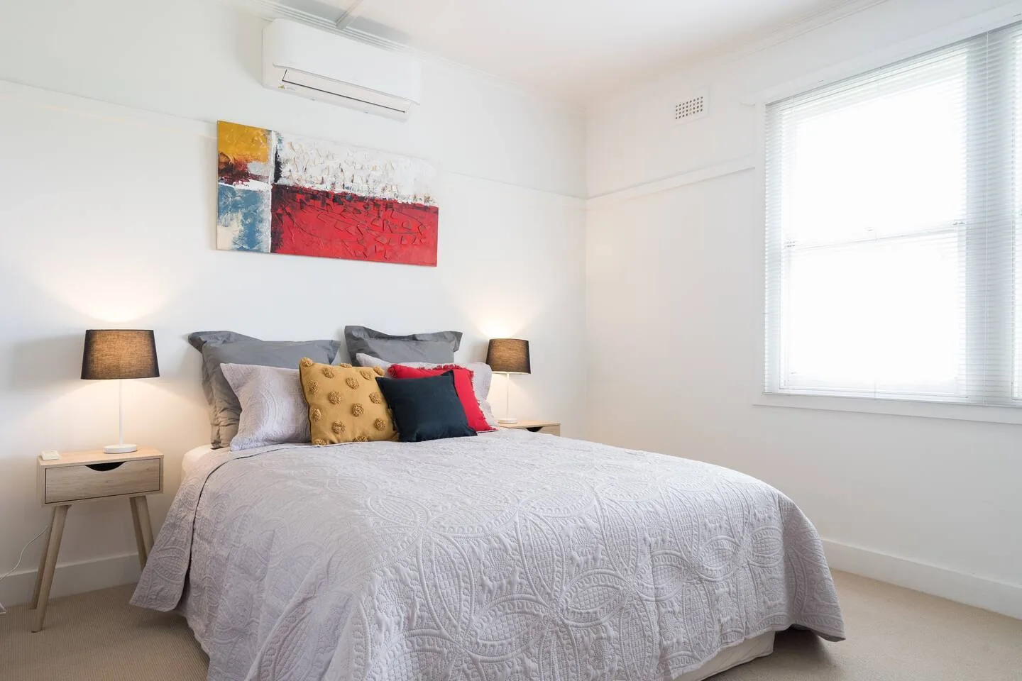 Un dormitorio con una cama, arte colorido en la pared y una ventana con persianas.