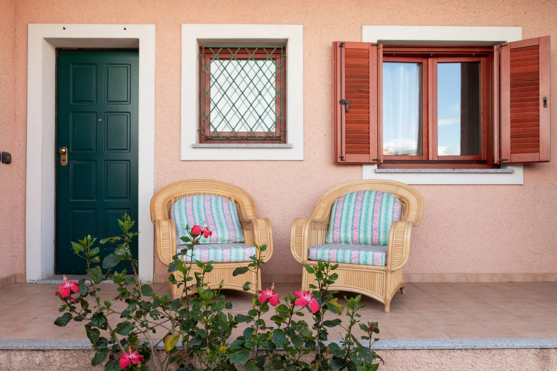 Una pared exterior rosa con una puerta oscura, una ventana abierta y dos asientos al aire libre.
