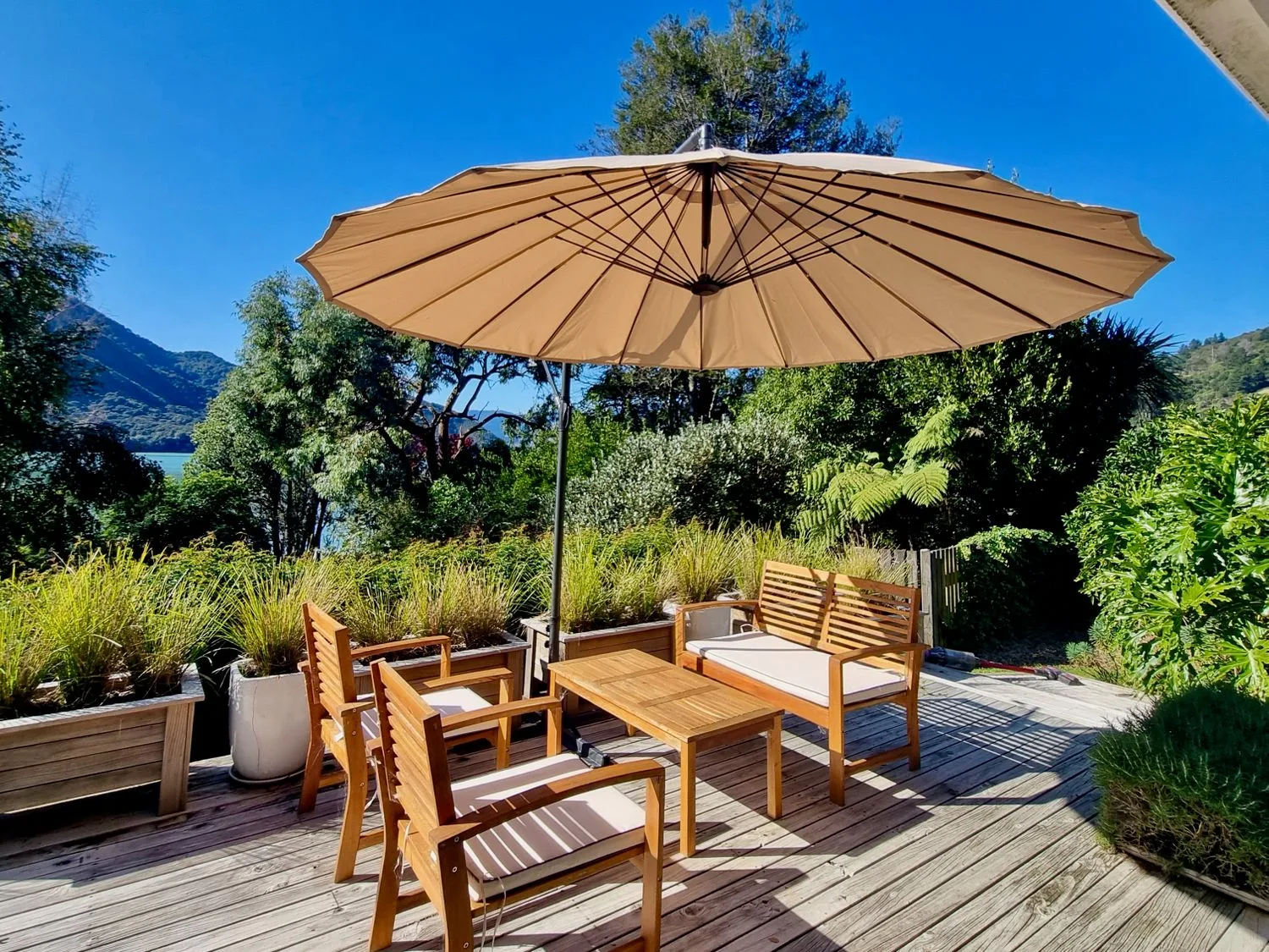 An outdoor wooden deck with chairs a bench a table and a large umbrella surrounded by plants at Pukenui Paradise - Havelock Holiday Home in Havelock
