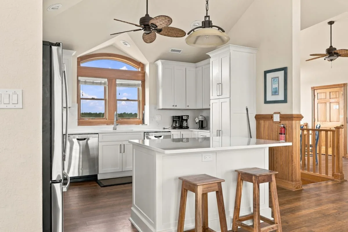 Una cocina luminosa con gabinetes blancos, una isla central y ventanas con marcos de madera.