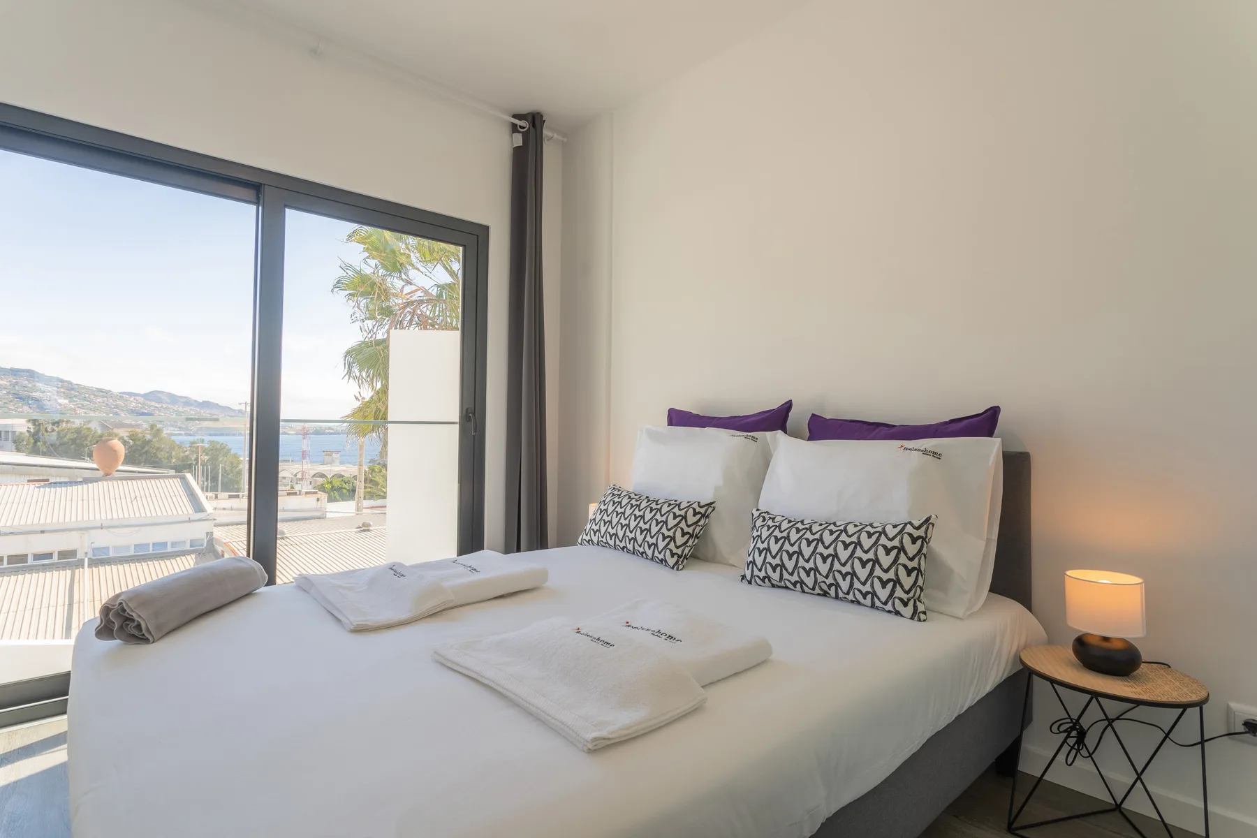 Un dormitorio con una cama bien arreglada junto a grandes ventanas con vistas a la costa.