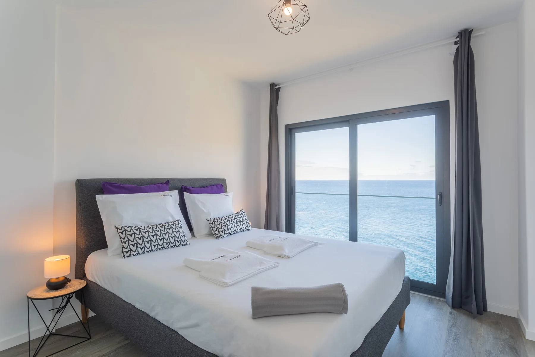 Un dormitorio con una cama junto a una ventana amplia con vistas al océano.