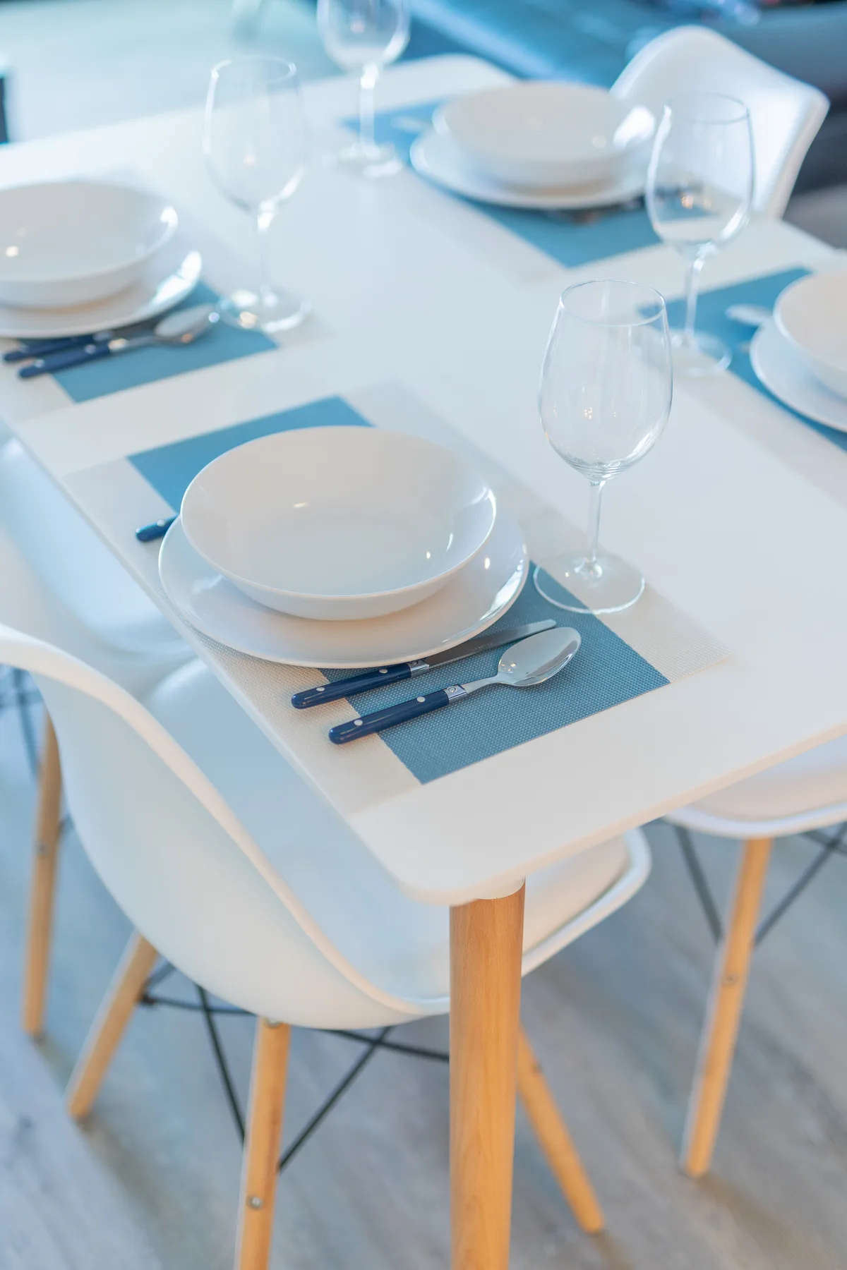 Una mesa de comedor está puesta con vajilla blanca, cristalería y manteles individuales azules.