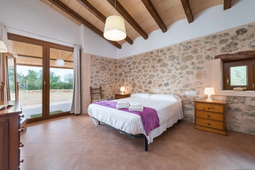 A bedroom with a large bed, exposed ceiling beams, and a stone wall.