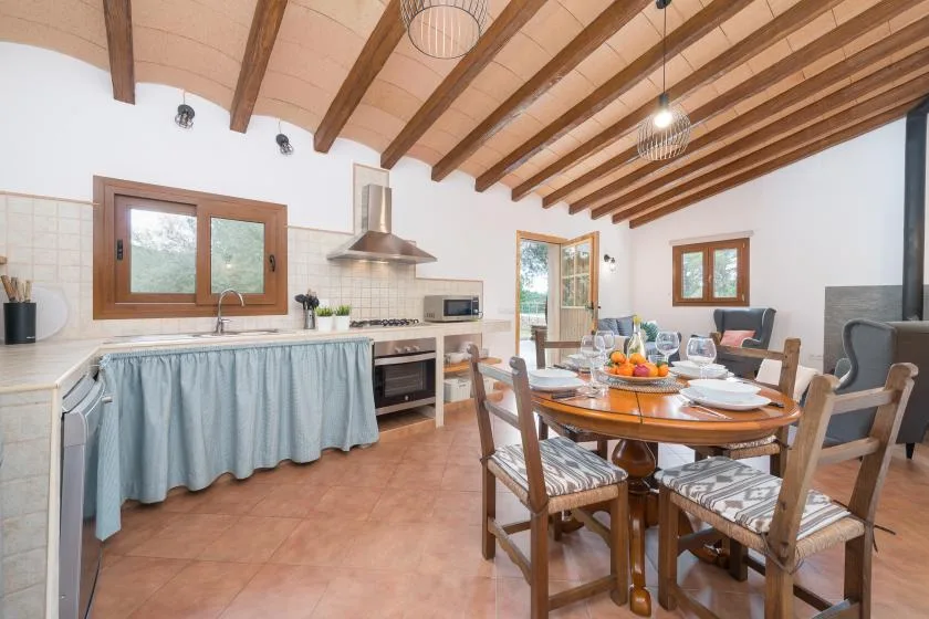 A dining area with a set table sits beside a kitchen under exposed wooden ceiling beams.