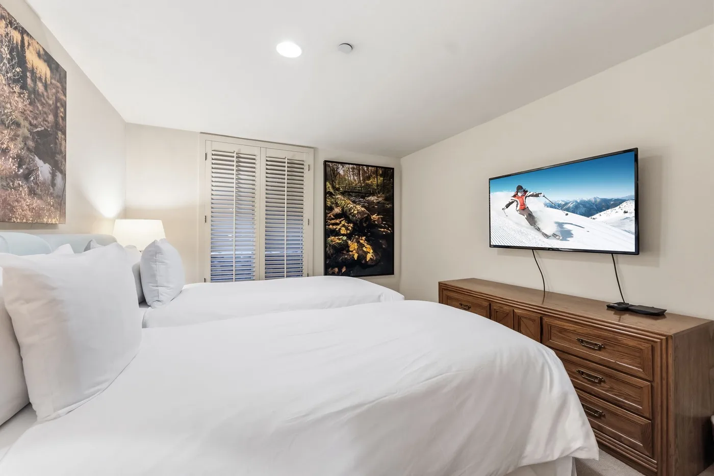 Un dormitorio luminoso con ropa de cama blanca, ventanas con persianas y un televisor en la pared.