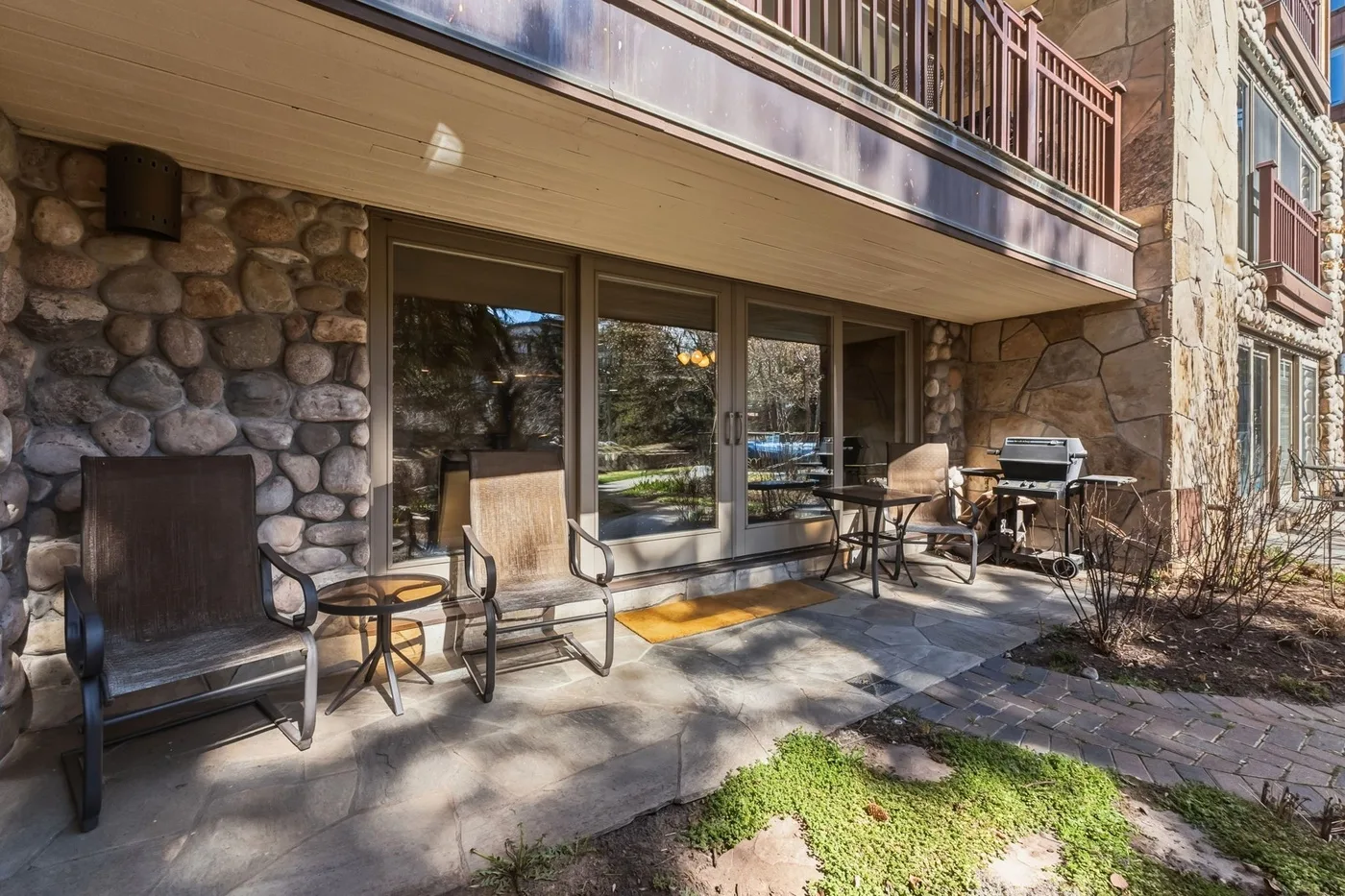 Un patio de piedra con asientos al aire libre y una parrilla junto a grandes ventanas.