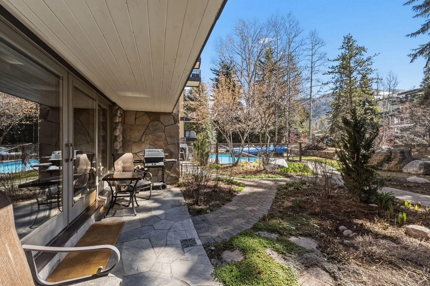 A paved patio with outdoor seating overlooks a landscaped garden and swimming pool.