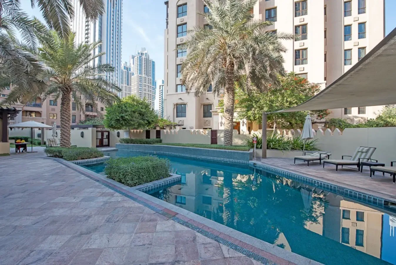 An outdoor swimming pool is surrounded by palm trees and nearby buildings.