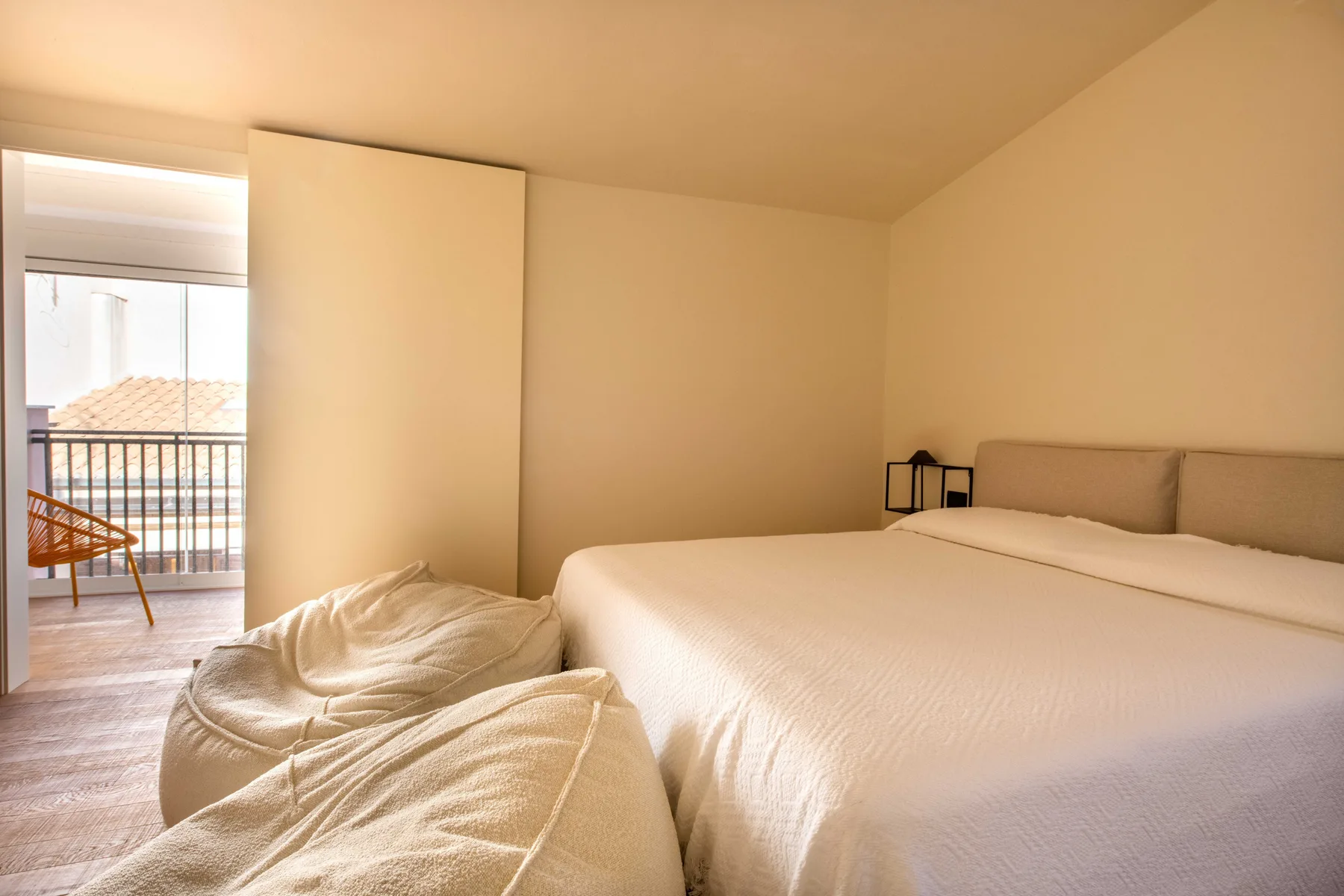 A bright bedroom with a large bed and a cushioned seat beside the open balcony doorway.