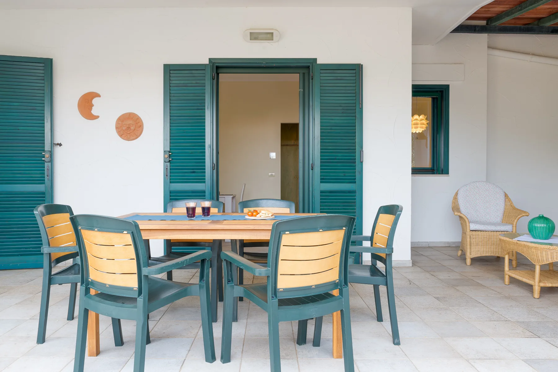A covered patio with a dining table and chairs beside open shuttered doors.