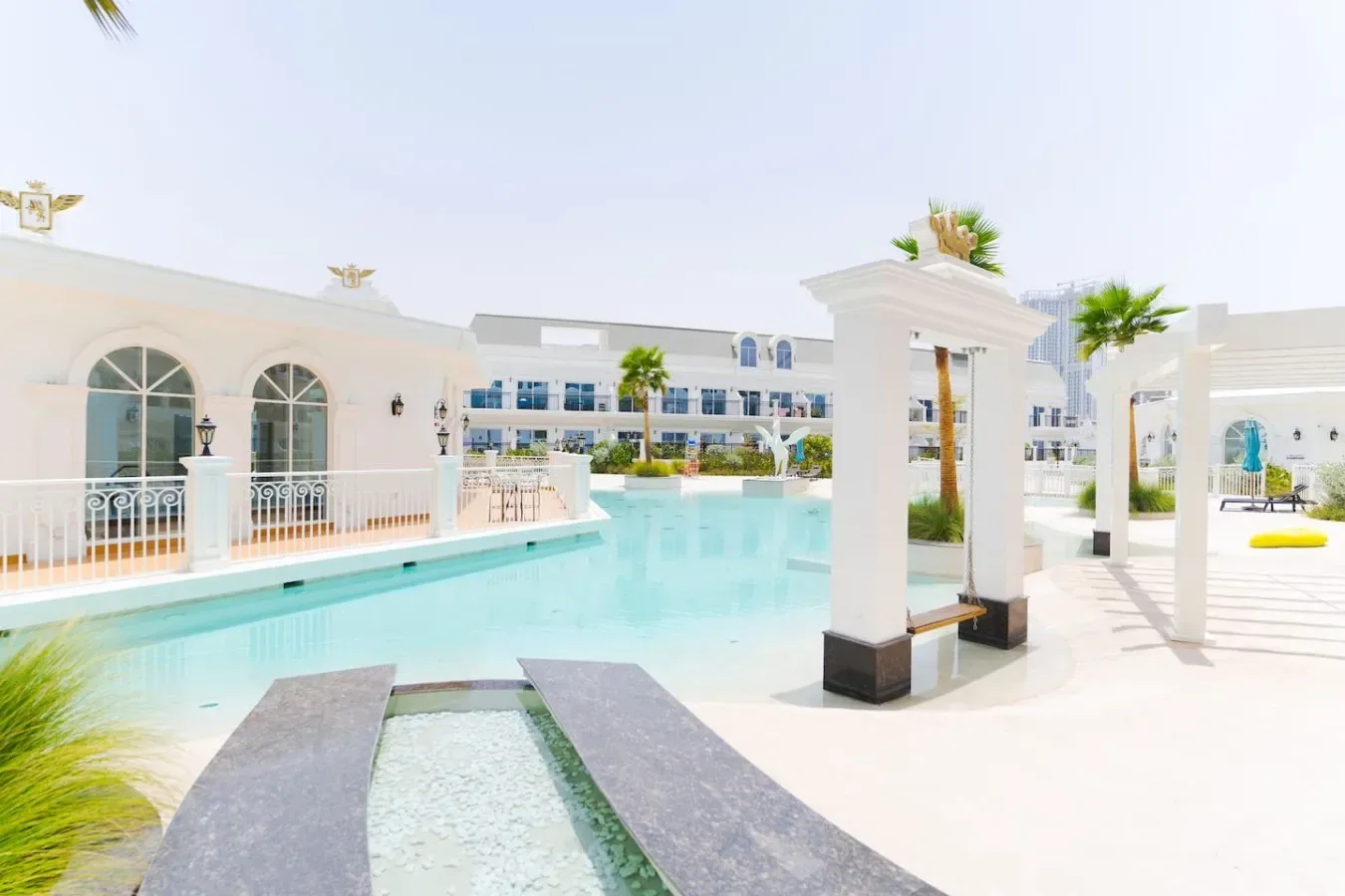 An outdoor pool area with white buildings and a pergola beside the water.