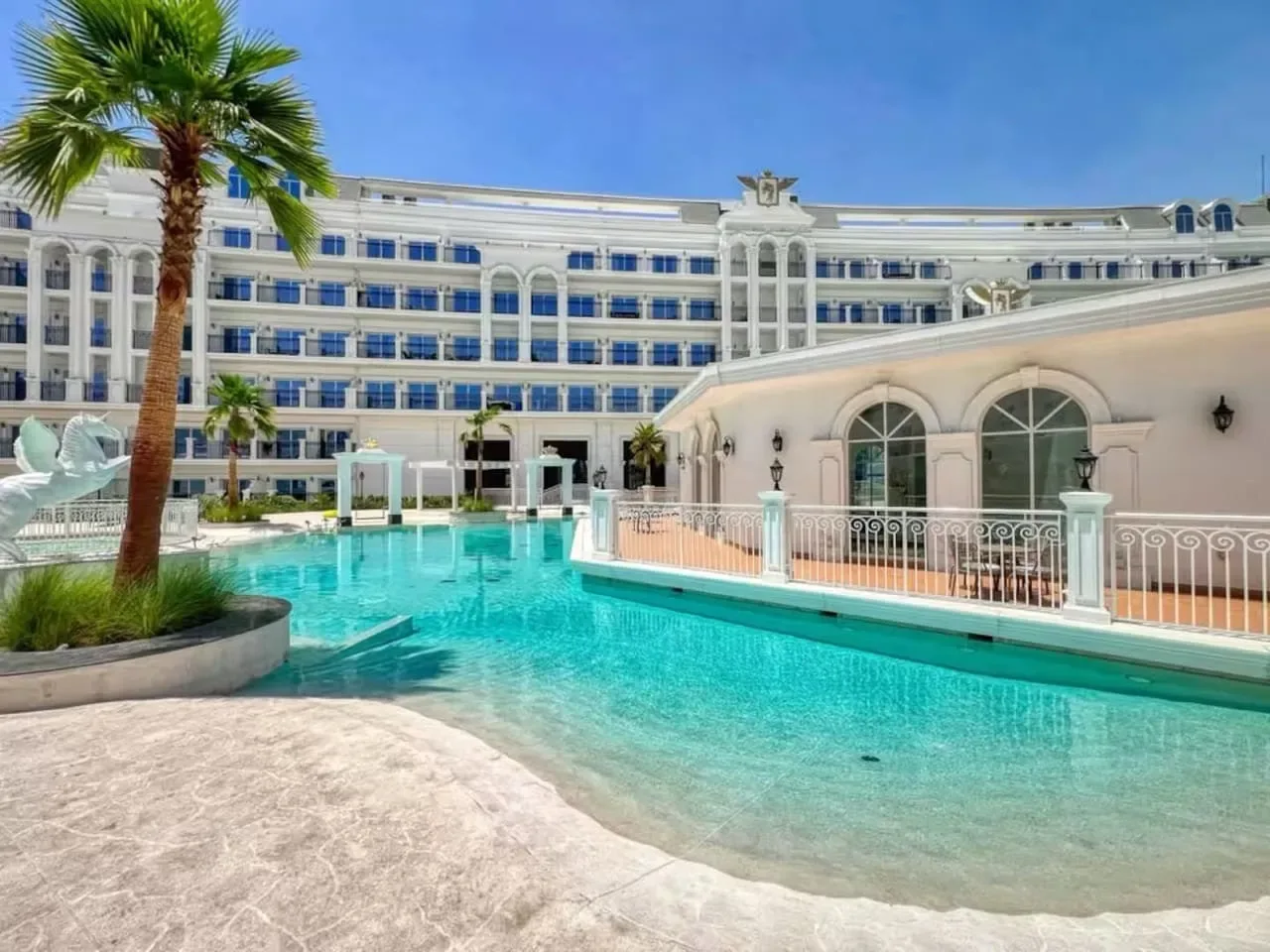 Una piscina turquesa al aire libre está junto a un edificio blanco y una palmera.