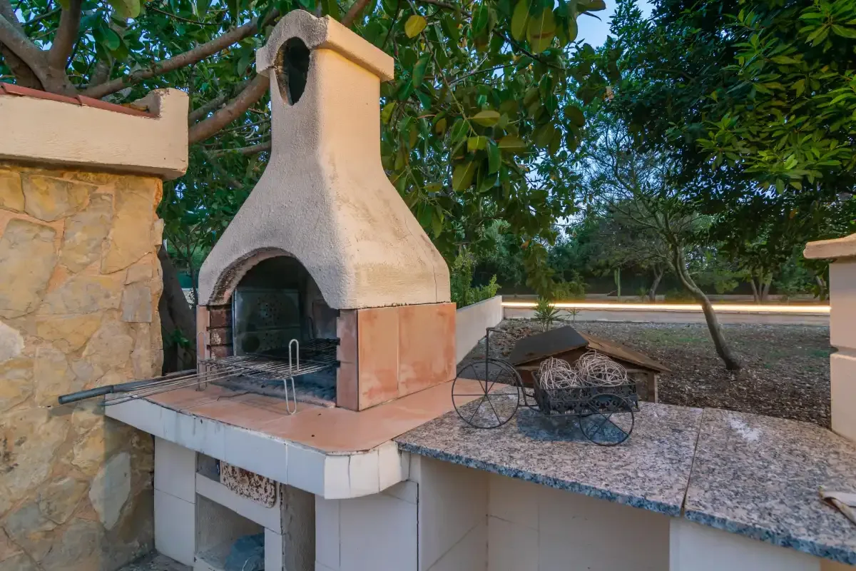 An outdoor masonry barbecue stands beside a stone countertop in a leafy garden.