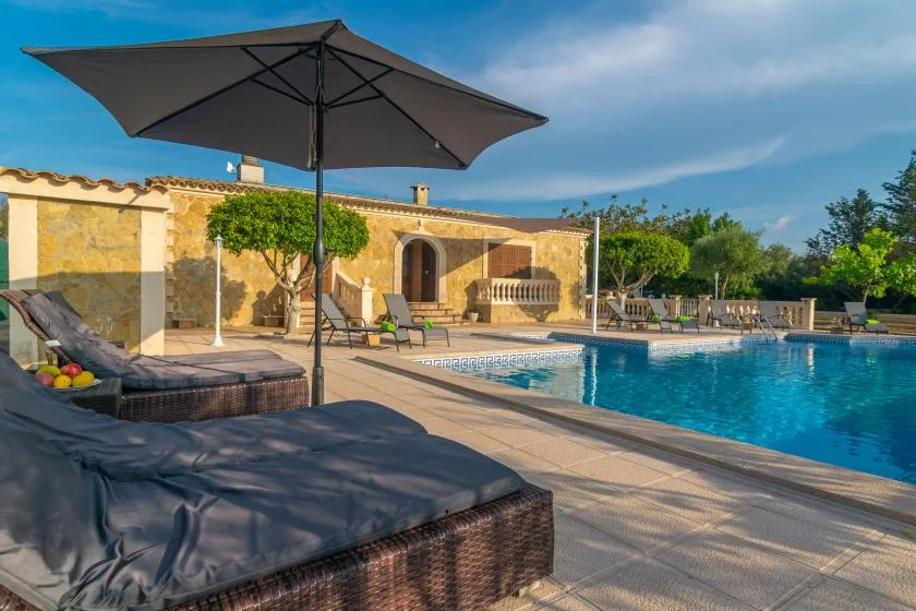 An outdoor swimming pool beside a stone house, with shaded seating nearby.