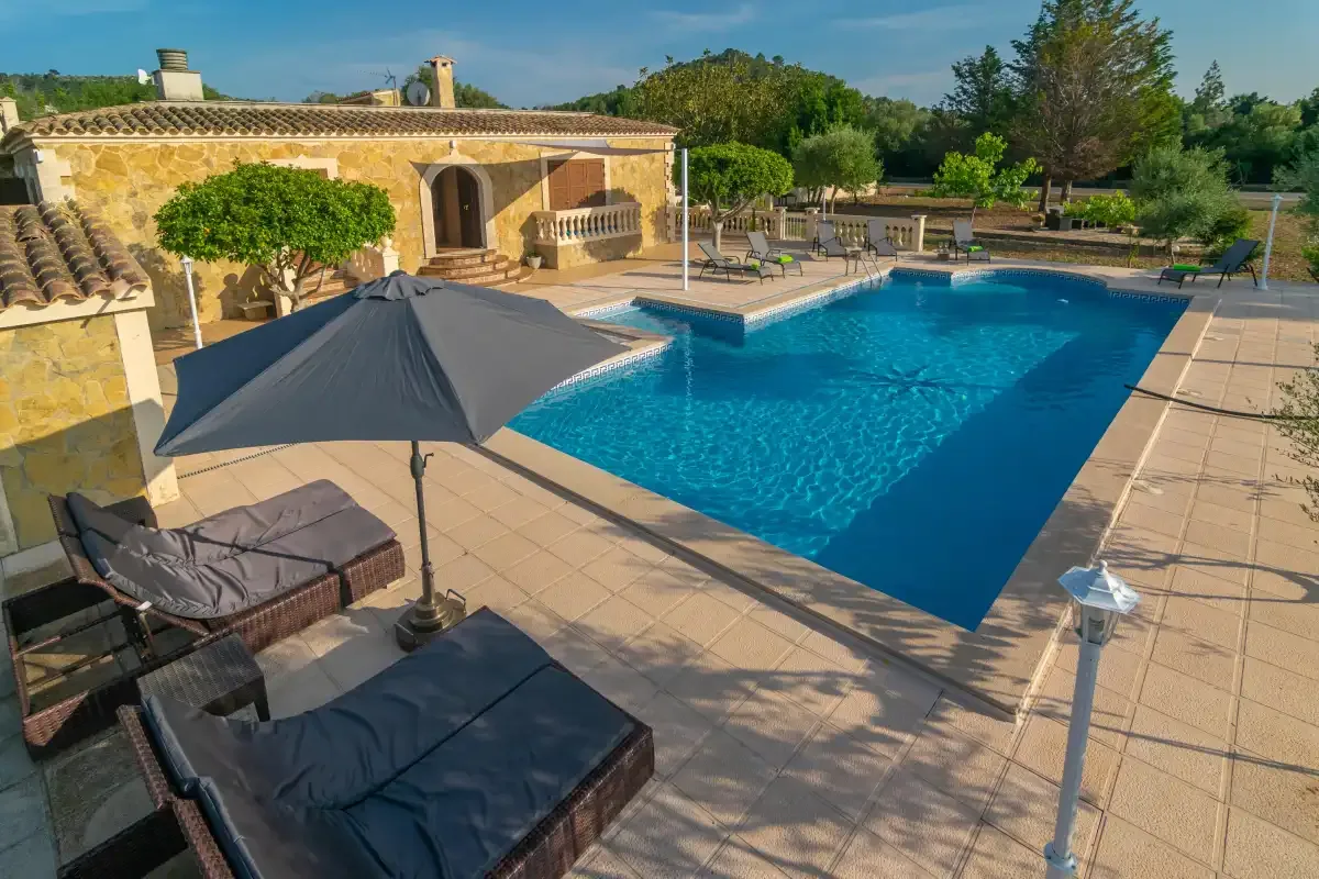 An outdoor swimming pool beside a stone house, with shaded lounge seating.
