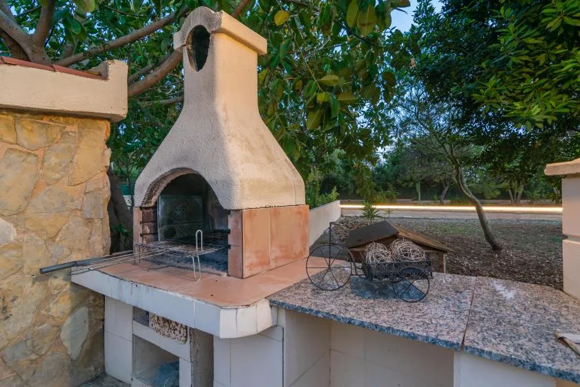 An outdoor masonry barbecue area sits beneath leafy trees.